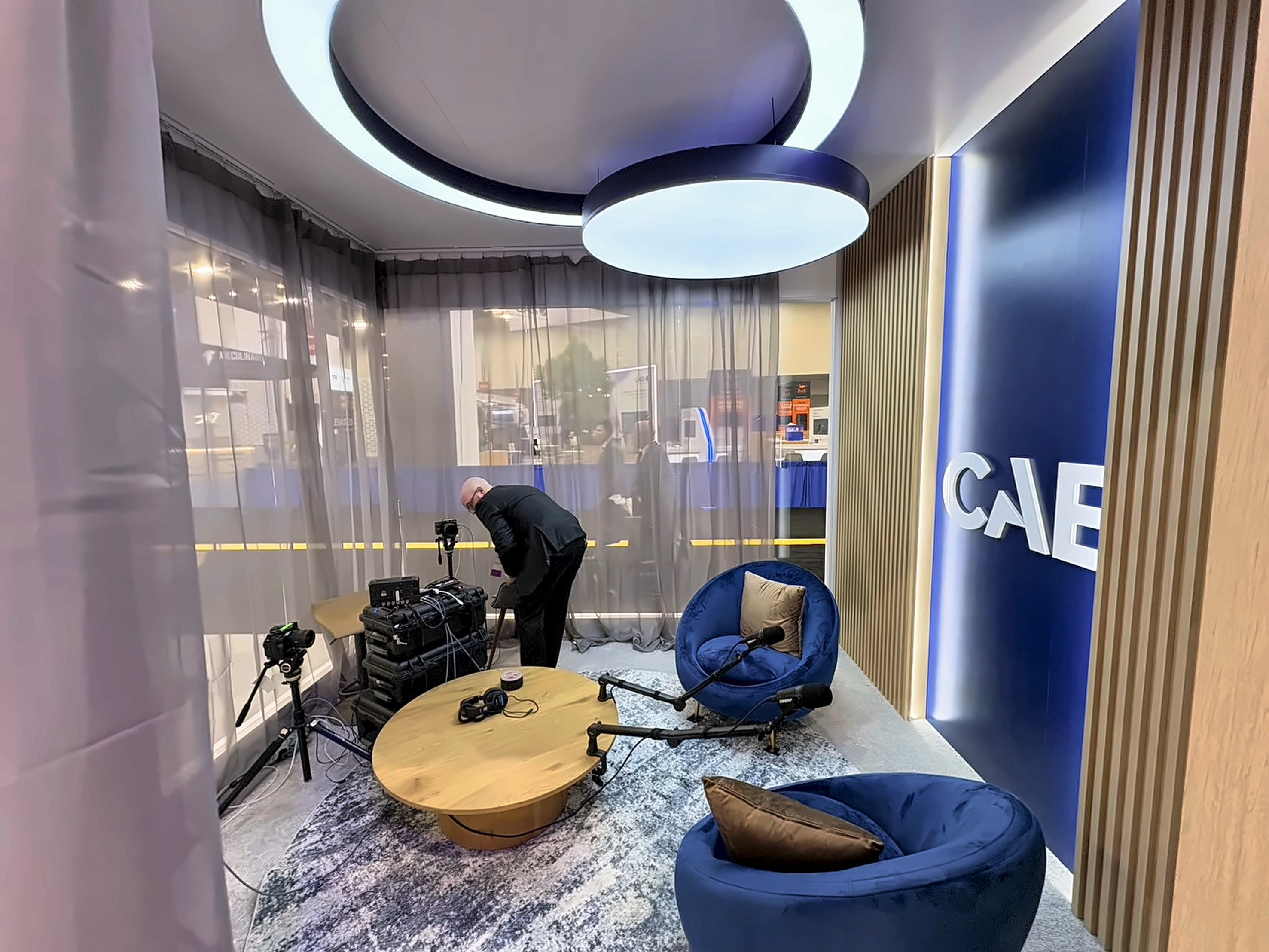 Podcast room behind sheer curtains with two ring lights, navy tub chairs, a round wood table, and a camera on a tripod