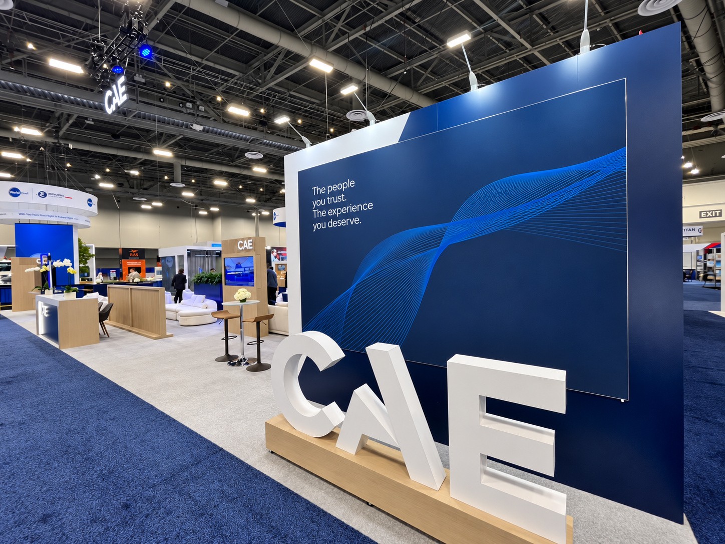 Large white CAE letters on a wood plinth in front of a navy wall with a blue wave graphic.